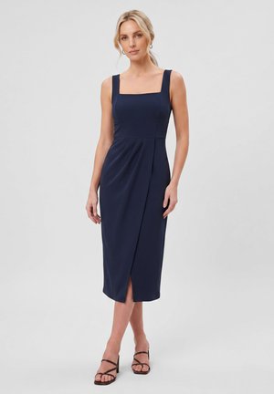 Frau mit hellem Haar in einem marineblauen ärmellosen Midikleid mit quadratischem Ausschnitt und vorderem Schlitz, trägt schwarze Riemchenabsätze, steht vor grauem Hintergrund.