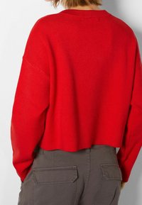 Roter, verkürzter Pullover aus Strickstoff mit Rundhalsausschnitt und überschnittenen Schultern, kombiniert mit grauen Cargo-Hosen, die mit Seitentaschen ausgestattet sind.