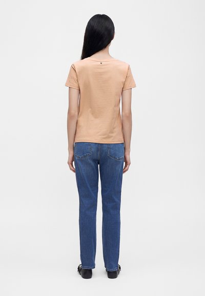 Femme aux longs cheveux noirs portant une chemise beige manches courtes, un jean bleu et des chaussures noires, debout de dos sur un fond uni.