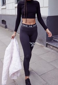 Musta pitkähihaine cropped paita ja korkeavyötäröiset mustat leggingsit, joissa on painettu brändäys. Kevyt tilkkutäkki takki pidettynä yhdessä kädessä. Mustat kengät.