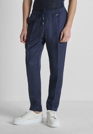 Persona que lleva puestos pantalones azul marino con cordón y bolsillos laterales, y zapatillas blancas informales, de pie frente a un fondo claro y sencillo.