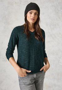 Maglione a coste di un verde scuro, con scollatura a V e maniche lunghe, presenta una leggera texture e una vestibilità rilassata. Indossato con jeans grigi chiari.