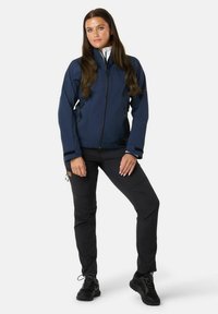Navy softshell jacket with a high collar, front zip, two zippered pockets, and black accents; paired with black pants and rugged shoes.