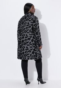 Grå djurmönstrad kappa med svarta leopardfläckar, långa ärmar och figursydd passform. Tyget verkar mjukt och texturerat. Bärs med svarta leggings och klackar.