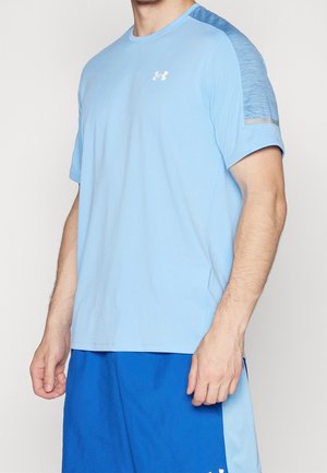 Mężczyzna w jasnoniebieskiej sportowej koszulce z krótkim rękawem z siateczkowymi panelami na ramionach i niebieskich sportowych szortach na jednolitym tle.