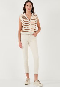Gebreide mouwloze vest in crème met bruine horizontale strepen, met een V-hals en grote knopen. Gecombineerd met off-white skinny jeans en beige sneakers.