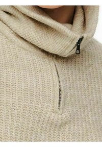 Beige gebreide trui met een hoge funnelneck, met een korte rits aan de voorkant. De textuur is zacht met een licht chunkyzak. 