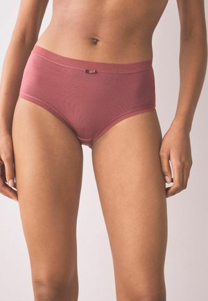 Žena stojaca pred jednoduchým pozadím, oblečená v bavlnených nohavičkách stredného pásu v odtieni ružovej s malou mašličkou na páse.