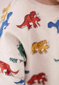 Sweatshirt beige avec des motifs de dinosaures colorés en bleu, rouge, vert et jaune. Matériau doux avec une coupe décontractée.