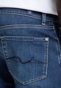 Mørkeblå denimjeans har en baklomme med en sydd bølgedetalj og et merke med teksten "for all mankind." Klassisk femlommers design.
