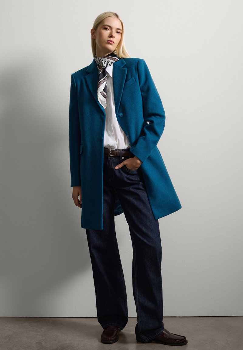 Een turquoise wollen jas over een witte shirt, gecombineerd met donkerblauwe denim jeans. Een gestreepte sjaal is om de hals geknoopt, wat de look compleet maakt.