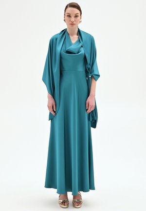 Donna che indossa un abito teal lungo fino a terra con scialle abbinato e sandali dorati con punta aperta, in piedi contro uno sfondo bianco semplice.