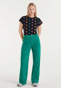 Camiseta negra con patrón de corazones multicolores, combinada con pantalones verdes de tiro alto. Ambos artículos presentan un ajuste casual y una textura suave.