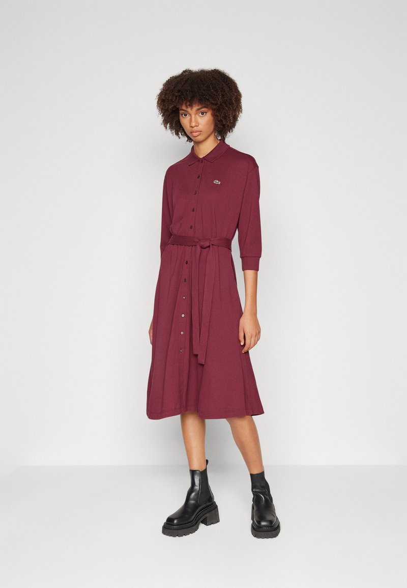 Bordeauxfarbenes, knielanges Kleid mit Kragen, Knopfverschluss vorne und tailliertem Gürtel. Hat drei Viertel lange Ärmel und ein Logo-Emblem.