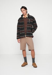 Fleecejacke mit grauen, roten und braunen Streifen, braunem Pullover, beigen Shorts, grauen Socken und braunen Schuhen. Modell steht vor einem schlichten Hintergrund.
