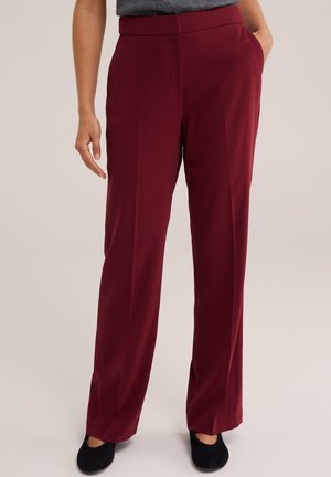 Femme portant un pantalon bordeaux taille haute à jambes droites avec poches latérales et chaussures noires, debout devant un fond uni.