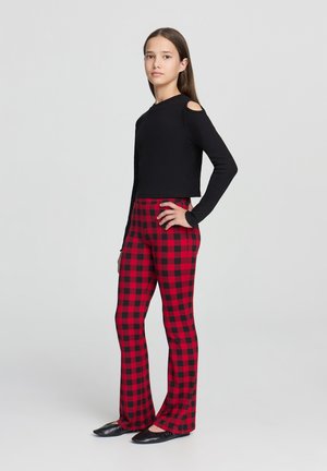 Maglietta nera a maniche lunghe con tagli, abbinata a pantaloni a palazzo a quadri rossi e neri. Texture morbide con design aderente, indossata con ballerine nere.