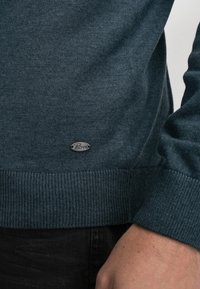 Sweter z dzianiny w ciemnym odcieniu teal z prążkowanymi mankietami i dołem, z małą srebrną łatą logo. Gładka textura, dopasowany krój.