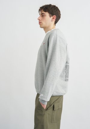 Jeune homme en sweat-shirt Nike gris avec inscription au dos et pantalon cargo vert olive, debout de profil les mains dans les poches.