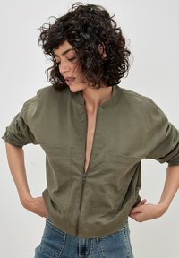 Olive green bomber jacket made of cotton, featuring a zip front, two chest pockets, and relaxed fit with rolled sleeves.