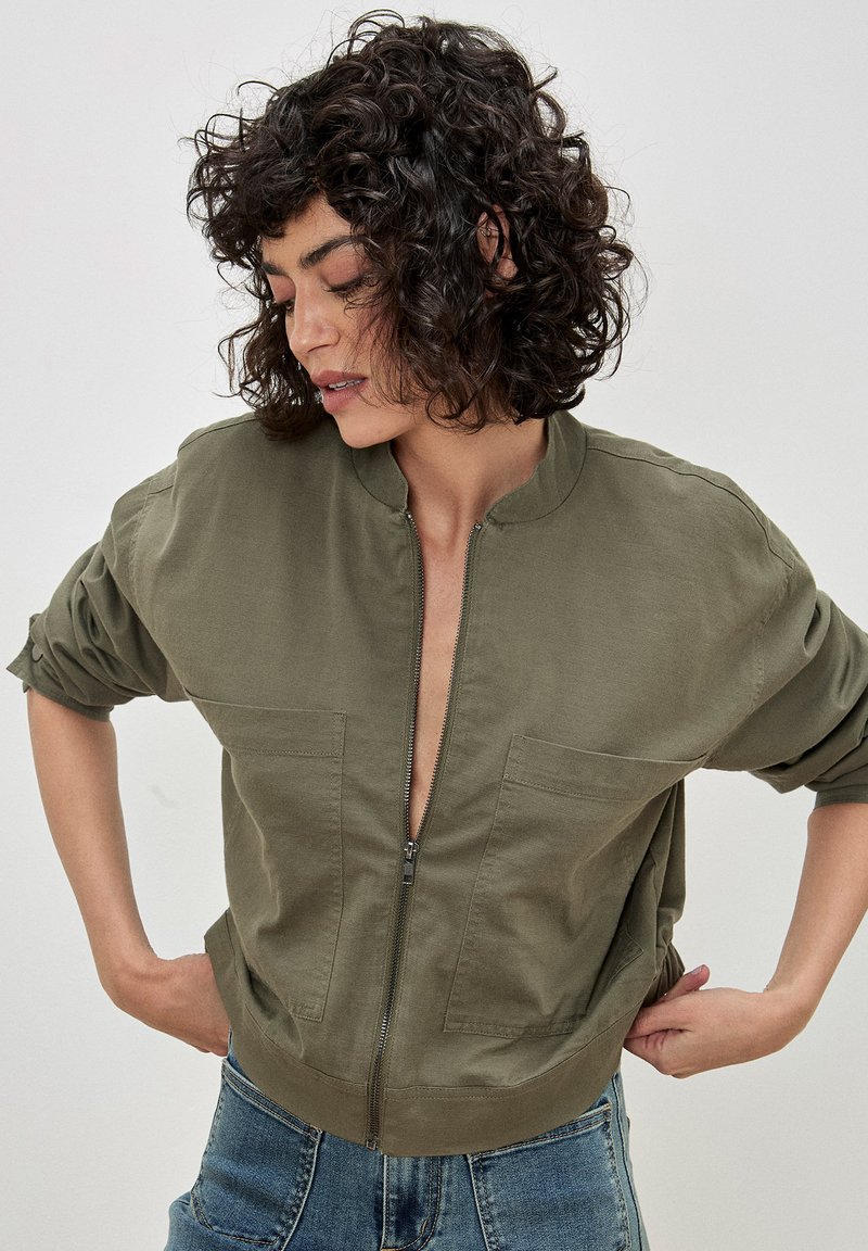 Olive green bomber jacket made of cotton, featuring a zip front, two chest pockets, and relaxed fit with rolled sleeves.