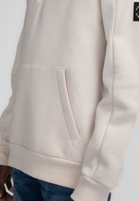 Lichtbeige sweatshirt met een kangoeroezak, geribbelde zoom en cuffed mouwen. Soepele textuur, minimaal ontwerp en een subtiel logo op de schouder.