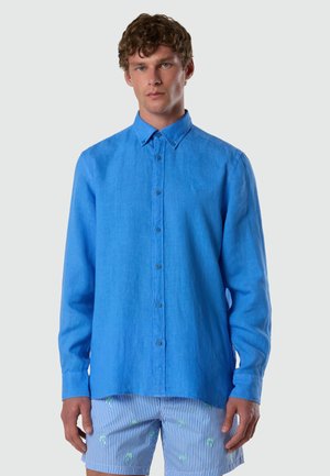 Uomo con capelli ricci che indossa una camicia azzurra con bottoni e pantaloncini a righe blu e bianche con piccoli motivi di palme verdi.