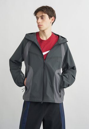 Νέος άνδρας που φορά γκρι φούτερ με κουκούλα πάνω από κόκκινο πουκάμισο με λευκό λογότυπο, στέκεται με τα χέρια στις τσέπες, κοιτάζοντας προς τα πλάγια.