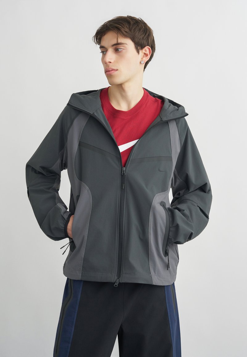 Junger Mann, der eine graue Kapuzenjacke über einem roten Shirt mit weißem Logo trägt, steht mit den Händen in den Taschen und schaut zur Seite.