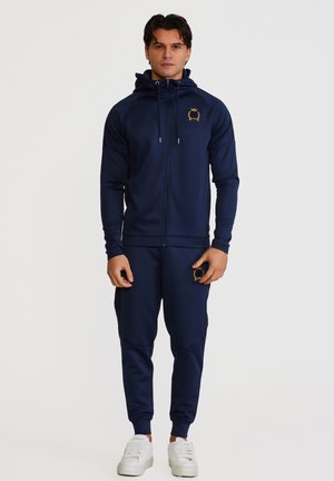 Mann står vendt fremover iført marineblå hettegenserdress med gull-emblem på bryst og lår, kombinert med hvite joggesko.