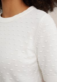Pull en tricot blanc avec un motif à pois texturé, col rond côtelé et manches courtes. Tissu léger avec une apparence douce et superposée.