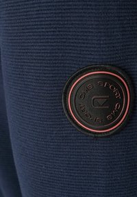 Granatowa tkanina z fakturą z wyraźną okrągłą naszywką w kolorze czarnym i czerwonym oznaczoną "CMS SPORT", z wypukłymi literami i różnymi fakturami.