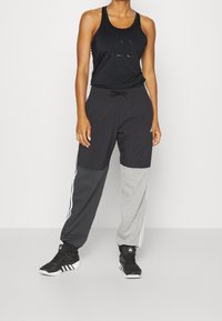Canotta nera senza maniche con logo, abbinata a pantaloni sportivi colorati in nero, grigio e grigio scuro. Scarpe da ginnastica nere.