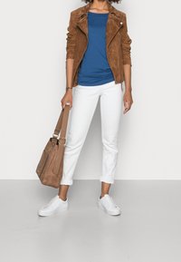 Giacca da motociclista in suede marrone, t-shirt blu, pantaloni bianchi con risvolto, e sneakers bianche; tiene una borsa marrone con tracolla regolabile.