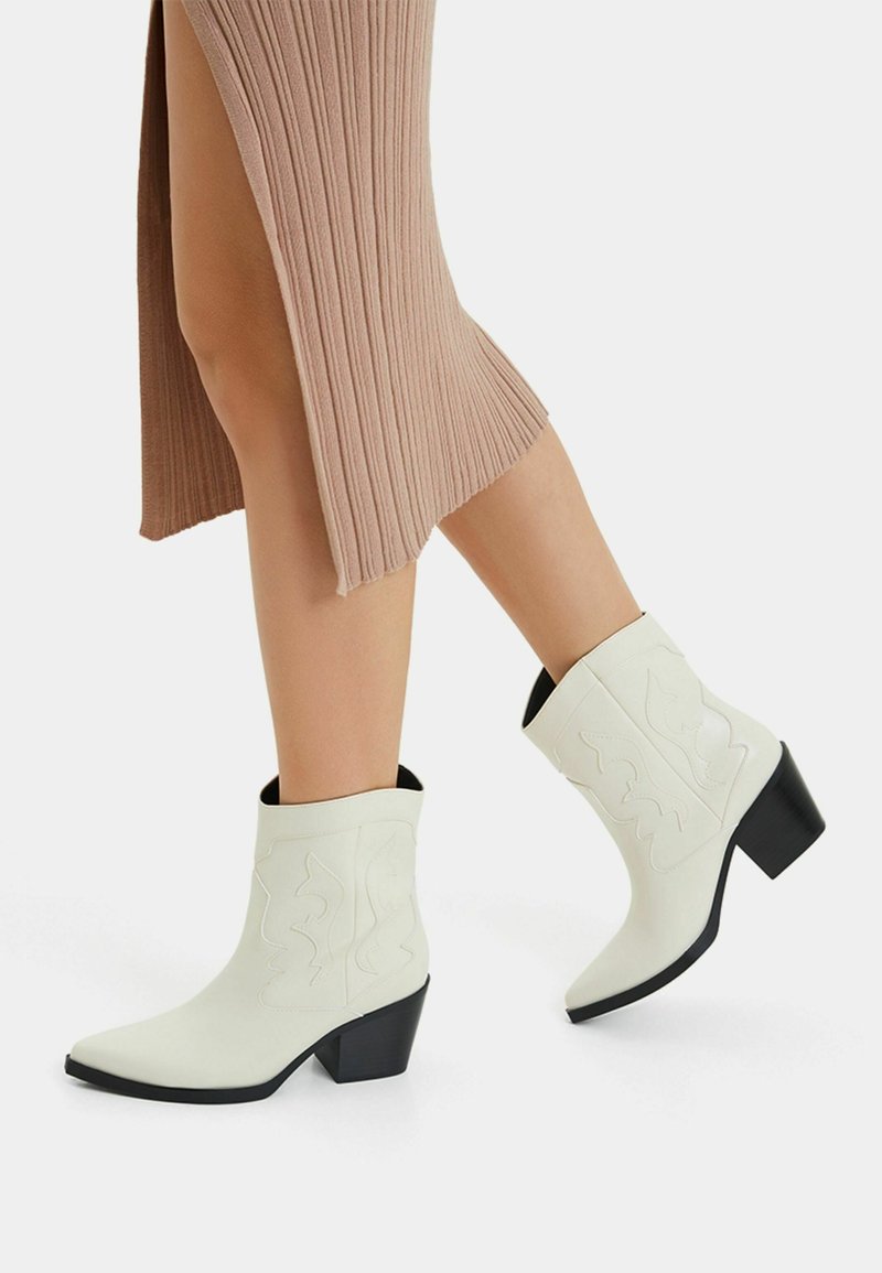 Białe skórzane kowbojskie buty z wydłużonym czubkiem, czarnym obcasem i tłoczonym wzorem, zestawione z prążkowaną spódnicą w kolorze taupe, która eksponuje nogi.