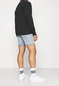 Osoba v černém triku s dlouhým rukávem, světle modrých džínových šortkách, bílých ponožkách s černými pruhy a bílých teniskách stojí zády.