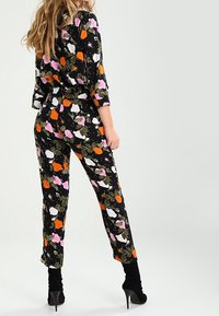 Combinaison florale avec un fond noir, ornée de grandes fleurs blanches, roses et orange, de feuilles vertes, et d'une coupe décontractée ; stylisée avec des talons hauts.