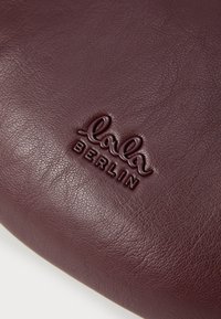 Βελούδινο δερμάτινο πουγκί σε μπορντό χρώμα με ανάγλυφο λογότυπο "lola BERLIN", υφή και στρογγυλό σχήμα.