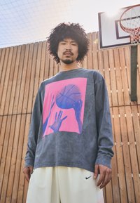Langermet grå t-skjorte med et rosa grafikk av hender og en basketball, sammen med hvite shorts. Trebakgrunn og basketballkurv synlig.