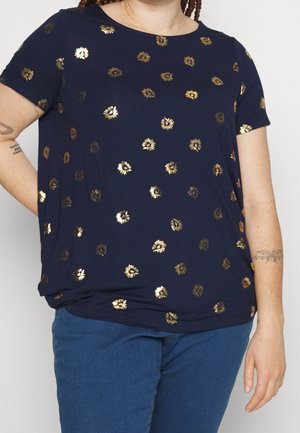 Osoba ubrana w granatową koszulkę z krótkim rękawem ozdobioną złotymi kwiatowymi wzorami oraz niebieskie spodnie, z widocznymi tatuażami na przedramionach.