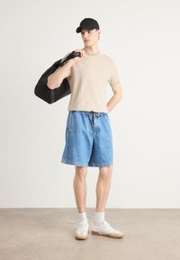 T-shirt beige à manches courtes, shorts en denim bleu, baskets blanches avec semelles en gomme et une casquette noire. Le modèle tient un sac noir.