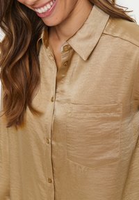 Gold satin shirt with a collar, button placket, and a chest pocket. The fabric has a smooth texture and a slight sheen.