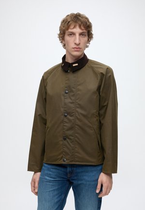 Junger Mann mit lockigem Haar, der eine olivgrüne, geknöpfte Jacke mit dunklem Kragen und blaue Jeans trägt und vor einem schlichten weißen Hintergrund steht.