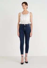 White sleeveless top with drawstring and ruffled hem, paired with dark blue high-waisted skinny jeans and black heels with polka dot accents.