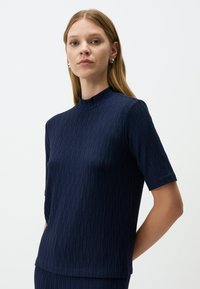 Navy blauwe top met korte mouwen en een hoge hals. Geweven stof met verticale ribbelpatronen, zachte stof en een ontspannen pasvorm.