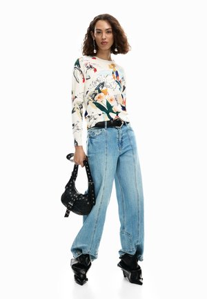 Woman stands wearing abstract floral print long-sleeve top, loose light blue jeans, black pointed boots, holding black studded handbag.
