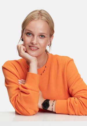 Bright orange sweatshirt with loose fit, paired with a black watch and chain bracelet. Hair is straight, and earrings are small and dark.
