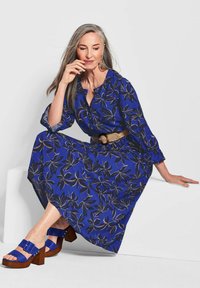 Blaues Blumenkleid mit langen Ärmeln und einem Taillengürtel. Klobige blaue Sandalen mit Absatz, Holzsohle und Schnallendetail.