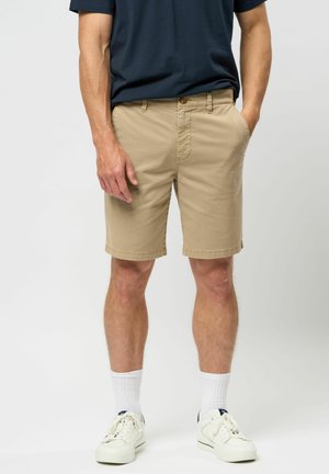 Homme portant un short beige longueur genoux, des chaussettes blanches, des baskets blanches et un t-shirt bleu foncé sur un fond clair uni.