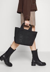 Borsa a spalla nera con logo DKNY, in tessuto resistente, con manici doppi e chiusura a zip, abbinata a una gonna a scacchi e stivali neri fino al ginocchio.